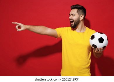 Displeased Angry Young Bearded Man Football Fan In Yellow T-shirt Cheer Up Support Favorite Team Hold In Hand Soccer Ball Point Finder Look Aside Isolated On Plain Dark Red Background Studio Portrait