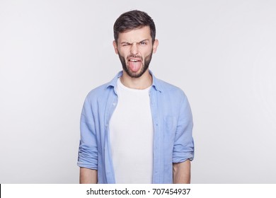 Displeased and bearded  young handsome blue eyed man employee wearing white shirt grimacing, making mouths, sticking out his tongue at camera trying to tease someone, acting like little naughty child.
