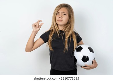 Displeased beautiful caucasian teen girl wearing sportswear holding a football ball over white wall shapes little hand sign demonstrates something not very big. Body language concept. - Shutterstock ID 2310771685