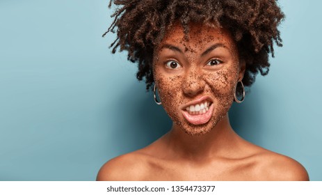 Displeased dark skinned woman frowns face with annoyance, doesnot want to make beauty procedures, has cleansing coffee mask on face, scrubs skin, stands over blue wall with empty space on left