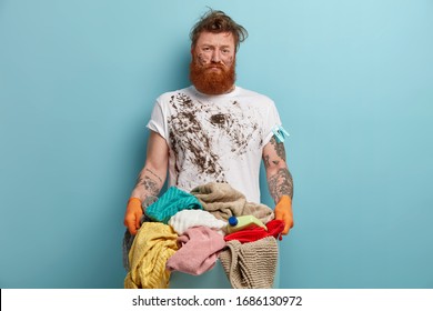 Displeased exhausted man holds basin with clean linen, finally finished laundry process, stands dirty after cleaning, has stressed look, poses against blue background. Housework, domestic chores