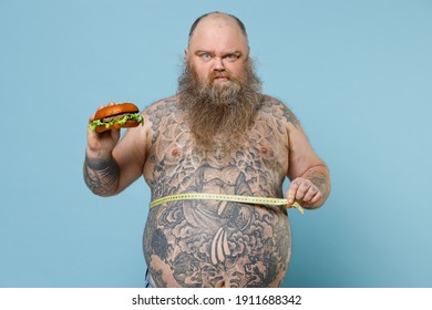 Displeased fat pudge obese chubby overweight man has tattooed naked big belly hold fast food burger measuring waist with tape isolated on blue background. Weight loss obesity unhealthy diet concept