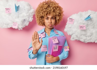 Displeased Female Student Makes Stop Gesture Asks Not To Bother Her Dressed In Blue Shirt Holds Folders Uses Sticky Notes For Writing Tasks To Do Poses Against Pink Background. Studying And Deadline