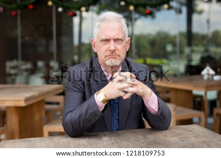 Displeased frowning senior businessman sitting at table in outdoor cafe. Angry bearded man scowling and looking at camera. Sullen concept