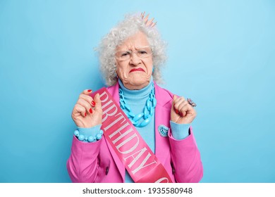 Displeased Grey Haired Woman Purses Lips Has Discontent Face Expression Raises Hands Prepares For Birthday Party Celebrates Anniversary Isolated Over Blue Background. Old People Emotions Concept