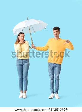 Displeased guy sharing umbrella with young woman on color background