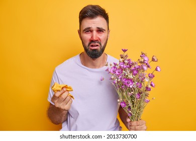 Displeased Ill Adult Man Suffers From Sinusitis Allergic Rhinitis Watery Red Eyes Holds Napkin Bouquet Of Wildflowers Isolated Over Yellow Wall Has Problems With Respiratory Tract. Seasonal Allergy