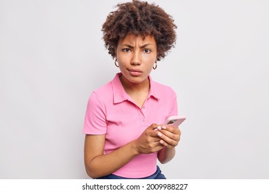 Displeased indignant dark skinned woman with curly hair uses mobile phone browses internet looks angrily at camera wears casual pink t shirt isolated over white background. Technology concept