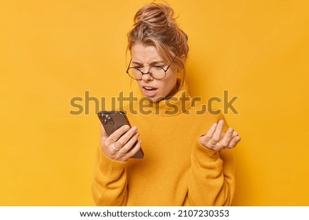Displeased indignant woman holds smartphone cannot understand how to use new application on device has disappointed face expression wears round spectacles and sweater isolated over yellow wall