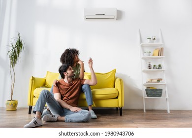 displeased interracial couple looking at air conditioner while sitting on sofa