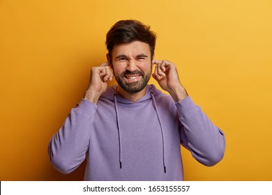 Displeased Irritated Man Plugs Ears, Cant Stand Loud Noise Or Sound, Ignores Conflict, Wears Violet Hoodie, Isolated On Yellow Background. Body Language Concept. Youngster Doesnt Want To Hear Music