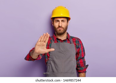 Displeased Male Engineer Makes Rejection Gesture, Says No, Keeps Palm Outstretched At Camera, Refuses Doing Manual Work, Wears Hardhat, Casual Uniform, Isolated On Purple Wall. Serious Constructor