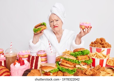 Displeased Mature Woman Holds Delicious Doughnut And Burger Has Unhealthy Eating Habits Eats Harmful Food Cannot Avoid Cosuming Cheat Meal Addicted To Sugar And Fat Dressed In Domestic Robe.