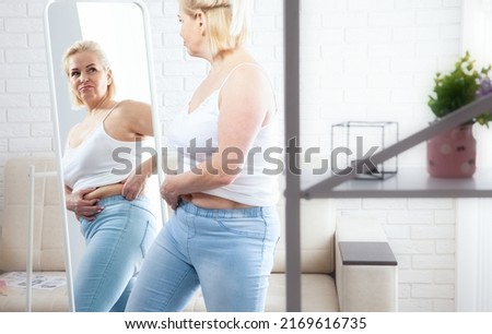 Displeased middle-aged woman looking at herself in the mirror and pinching stomach fat in domestic interior. Sad plus size woman looking her reflection in the mirror with sorrow at home.