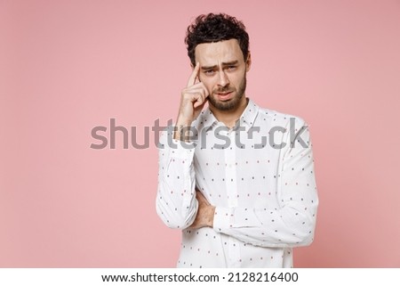 Displeased preoccupied worried puzzled young bearded man 20s wearing basic casual white shirt standing put hand on head looking camera isolated on pastel pink color wall background studio portrait