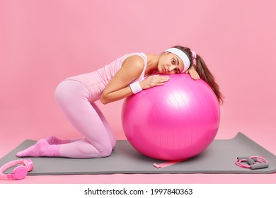 Displeased Sad Slim Woman Leans At Fitness Ball Has Tired Expression Trains At Home Gym Dressed In Activewear Poses On Knees Isolated Over Pink Background. People Sport And Gymnastics Concept