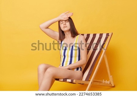 Displeased tired young woman girl wearing striped swimsuit sitting on deck chair isolated on yellow background, having summer vacation, putting hands on head, has sunstroke