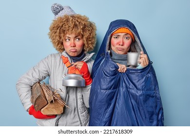 Displeased Two Female Hikers Have Camping Trip In Forest Drink Hot Beverage From Metal Cup Carry Wood Wear Warm Winter Clothes Wrapped In Sleeping Bag Isolated Over Blue Background. Hiking Concept