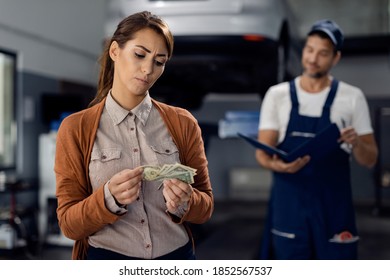 Displeased Woman Counting Money To Pay A Bill For Car Repair At Workshop.