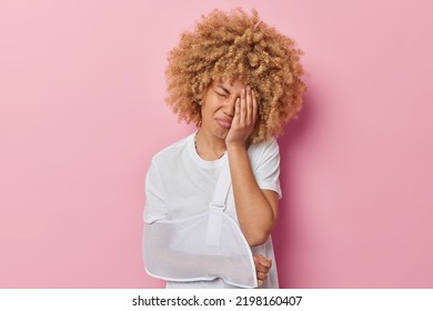 Displeased Woman With Curly Hair Covers Hand With Palm Feels Pain After Getting Into Accident Has Broken Arm Wears Sling Has Bone Fracture Dressed In Casual T Shirt Isolated Over Pink Background