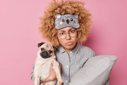 Displeased Woman With Curly Hair Dressed In Pajama And Sleepmask Holds Pillow Prepares For Sleep Holds Pug Dog Poses Against Pink Background Enjoys Company Of Best Friend. Friendship Concept