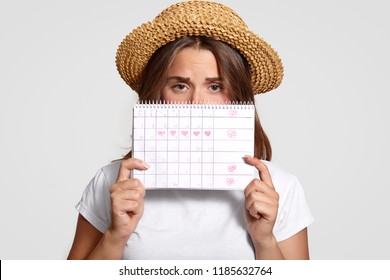 Displeased woman holds perids calendar, wears straw hat, has discontent expression, dressed in casual t shirt, has problems with womens health, isolated over white background. Menstruation concept