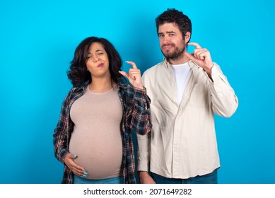 Displeased Young Couple Expecting A Baby Standing Against Blue Background Shapes Little Hand Sign Demonstrates Something Not Very Big. Body Language Concept.