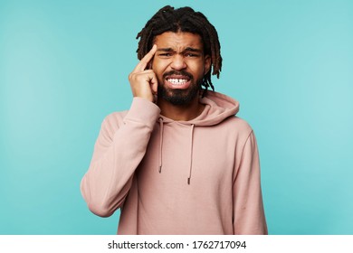 Displeased young dark haired bearded male grimacing his face and holding finger on temple while looking at camera, standing over blue background in casual wear - Shutterstock ID 1762717094
