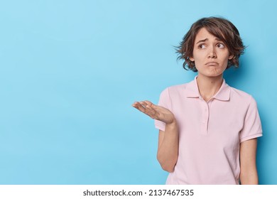 Displeased young European woman with trendy hairstyle looks upset raises palm has indignant sad expression wears casual t shirt concentrated away poses against blue background blank space away