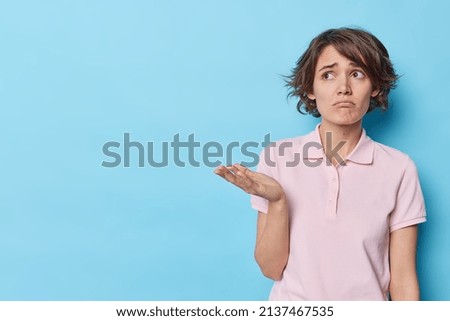 Displeased young European woman with trendy hairstyle looks upset raises palm has indignant sad expression wears casual t shirt concentrated away poses against blue background blank space away