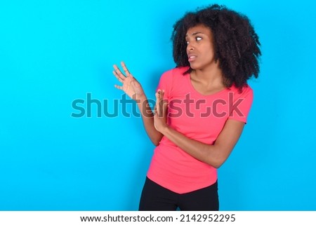 Displeased Young girl with afro hairstyle wearing pink t-shirt over blue background keeps hands towards empty space and asks not come closer sees something unpleasant