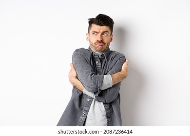 Displeased Young Man Feeling Discomfort, Hugging Himself And Staring Left At Something Embarrassing Or Exposing, Standing Uncomfortable On White Background