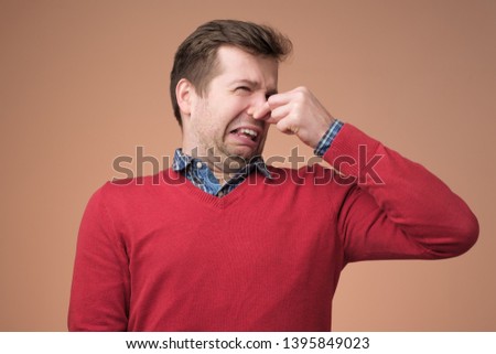 Displeased young man in red sweater plugs nose as smells something stink and unpleasant, feels aversion. Guy hates disgusting scent which comes from somewhere