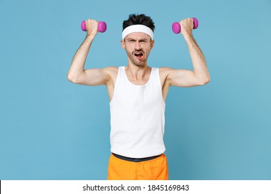 Displeased Young Strong Sporty Fitness Man With Skinny Body Sportsman In White Headband Shirt Shorts Doing Exercise With Dumbbells Isolated On Blue Background. Workout Gym Sport Motivation Concept