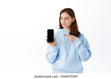 Displeased Young Woman Showing Bad Application On Her Phone, Pointing At Empty Smartphone Screen And Grimacing Upset, Standing Against White Background