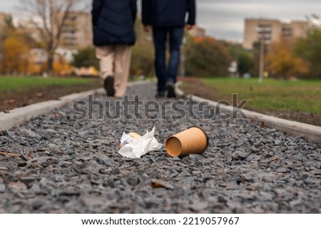 Disposable coffee cup and a piece of croissant thrown away on the ground  in park with young couple walking in the background. Environmental pollution and litter concept.