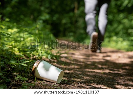 Disposable coffee cup thrown away on the ground in forest with person walking in the background. Environmental pollution and litter. Plastic waste and recycling problem