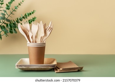 Disposable dishes and utensils against beige green background. Biodegradable wooden cutlery in brown paper cups on plates. Eco-friendly tableware and recycling concept. Copy space. Front view.