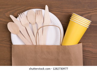 Disposable Paper Plates And Cups And Wooden Cutlery On Wooden Table In A Paper Bag