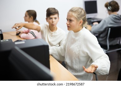 Dissapointed Teenager Girl Making Helpless Hand Gesture While She Using Computer In Classroom. Boy Sitting Next To Her And Pointing On Display.