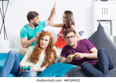 Dissapointed Young Couple With Gamepads Sitting On Large Bean Bags With The Other Couple Celebrating Victory In The Background