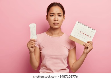 Dissatified Gloomy Woman Holds Menstruation Sanitary Soft Pad, Being In Bad Mood During Critical Days, Holds Periods Calendar With Marked Red Days, Wears Rosy T Shirt. Women Hygiene Concept.