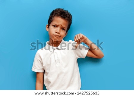 dissatisfied african american boy in white polo showing dislike on blue isolated background, sad 10 year old child rejects and disapproves and shows bad grade