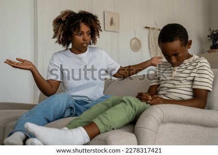 Dissatisfied African American woman mother scolding upset son for bad behavior while sitting together on sofa in living room. Disappointed parent mom talking with kid, disciplining child at home