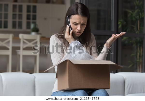 Dissatisfied angry woman, cheated client sit on\
sofa check received box, damaged or broken goods in parcel, talks\
to customers support, express complaints, looks annoyed. Bad\
delivery services\
concept
