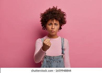 Dissatisfied annoyed woman with Afro hair smirks face and clenches fists, looks angrily at someone, promises to revenge or punish for bad behavior, has annoyed expression, poses over rosy background
