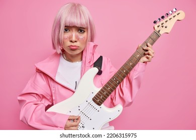 Dissatisfied Asian Woman With Pink Dyed Hair Plays Electic Acoustic Guitar Learns Playing Musical Instrument Dressed In Jacket Frowns Face Has Bad Mood Isolated Over Rosy Background. Upset Musician