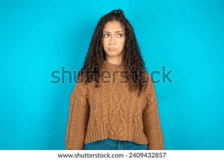 Dissatisfied Beautiful arab teen girl with curly hair wearing knitted sweater purses lips and has unhappy expression looks away stands offended. Depressed frustrated girl.