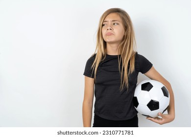 Dissatisfied beautiful caucasian teen girl wearing sportswear holding a football ball over white wall purses lips and has unhappy expression looks away stands offended. Depressed frustrated model. - Shutterstock ID 2310771781