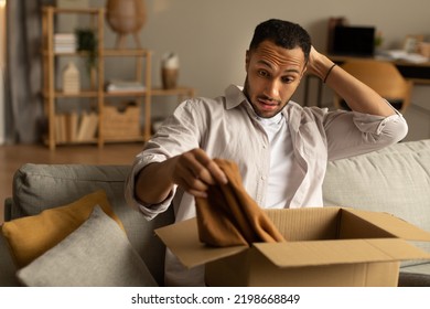 Dissatisfied Buyer Concept. Frustrated Black Man Unboxing Package After Delivery, Holding Clothes, Sitting On Couch At Home, Copy Space. African American Guy Unhappy About Online Purchase